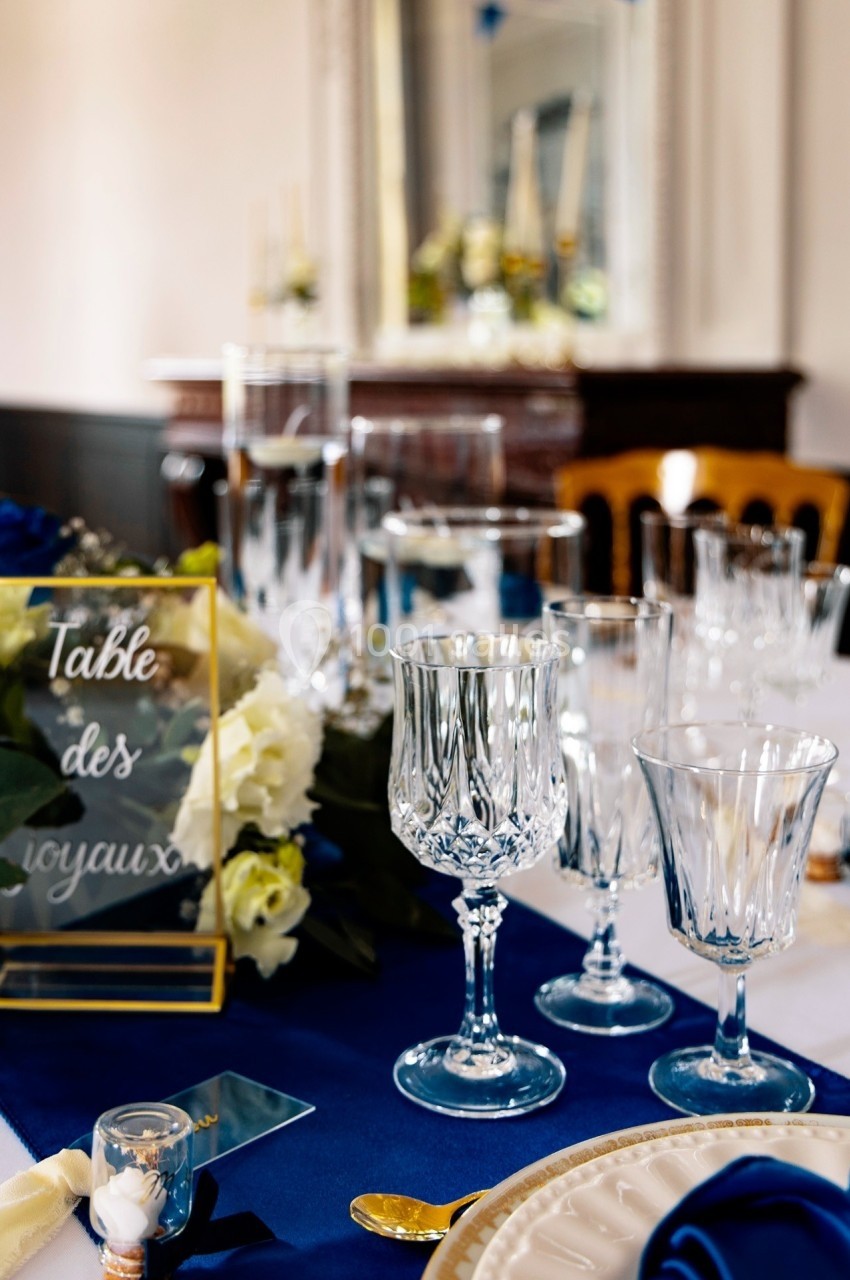 Table élégamment dressée avec nappes bleues, verres en cristal, fleurs blanches et panneau ’Table des voyaux’.