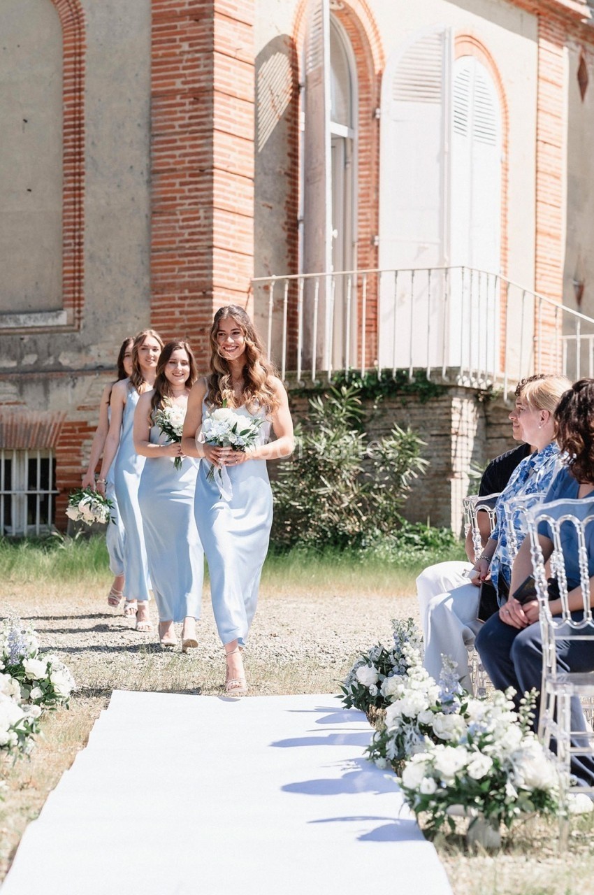 Des demoiselles d'honneur en robes bleu clair marchent sur une allée blanche lors d'une cérémonie en extérieur.