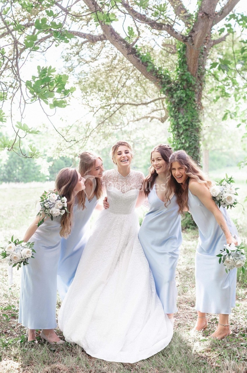 Une mariée souriante entourée de quatre demoiselles d'honneur en robes bleu clair dans un jardin verdoyant.