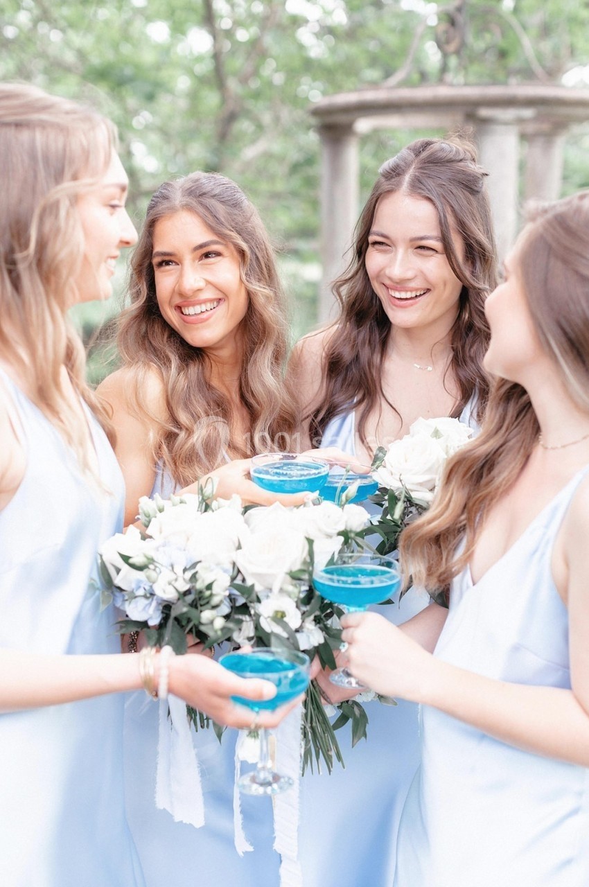Quatre jeunes femmes en robes pastel sourient et tiennent des cocktails bleus et un bouquet de fleurs blanches.