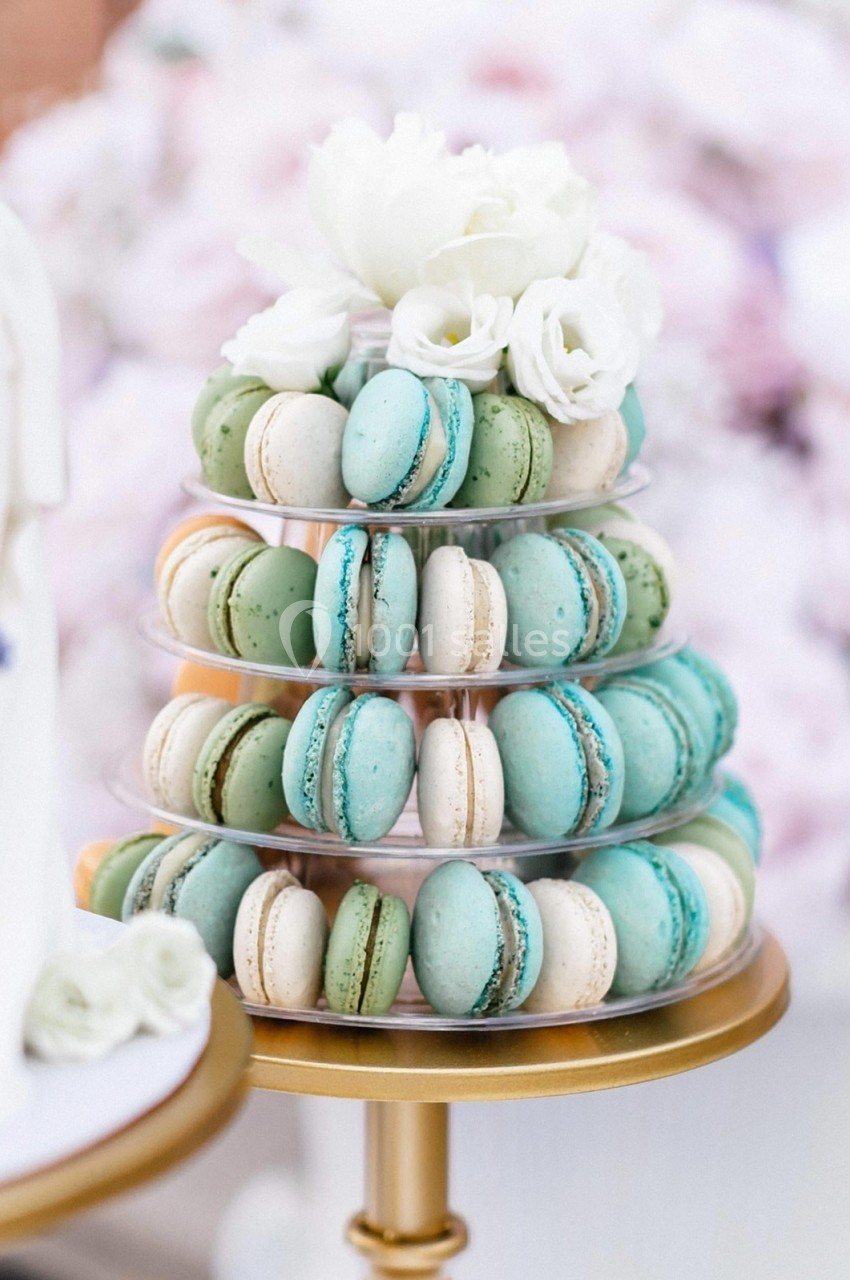 Tour de macarons pastel bleus et verts sur un présentoir doré, décorée de fleurs blanches en haut.