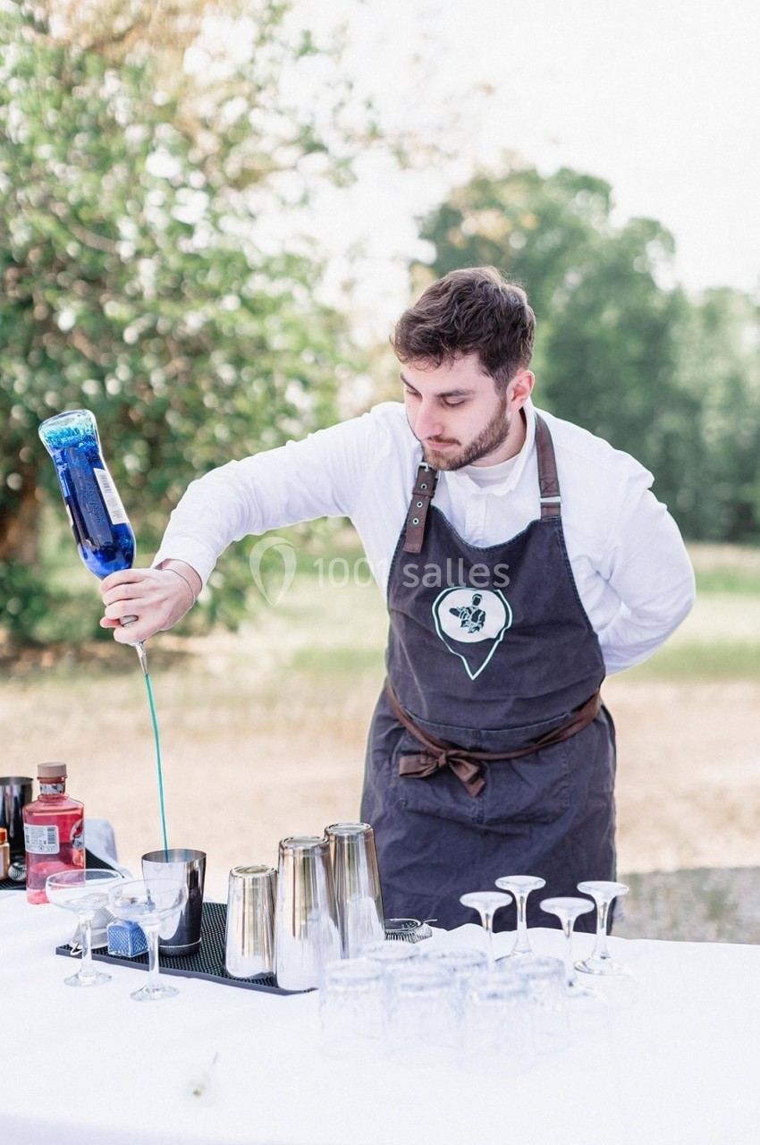 Un barman verse un liquide bleu dans un shaker sur une table dressée en extérieur.