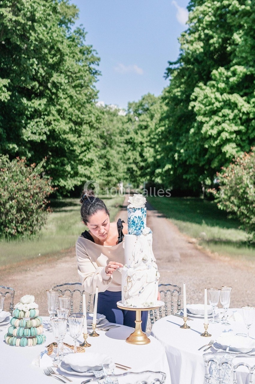 Une femme décore un gâteau à étages sur une table dressée en extérieur, entourée de verdure.