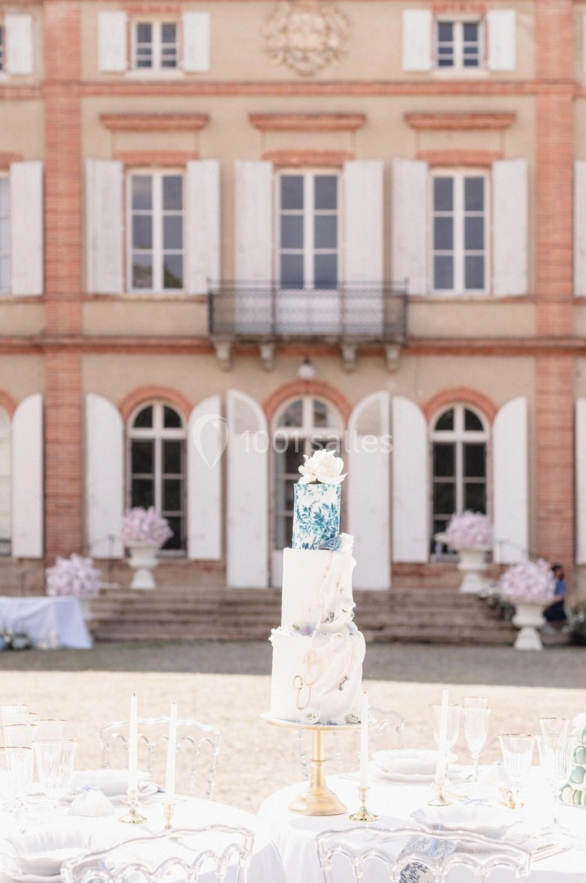 Gâteau de mariage à plusieurs étages décoré, placé sur une table en extérieur devant un bâtiment ancien avec des volets…