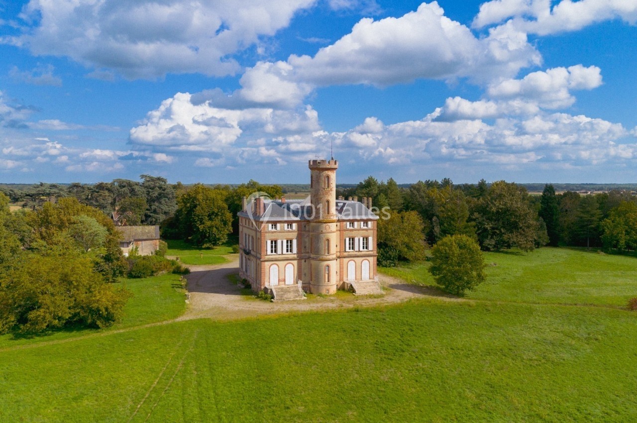 Château en briques avec une tour centrale, entouré de pelouses et d'arbres sous un ciel partiellement nuageux.