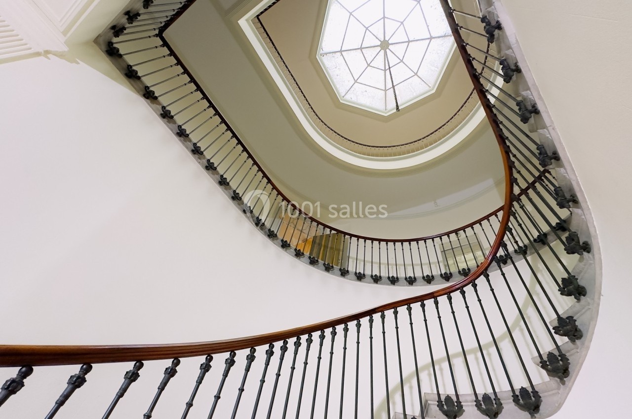 Escalier en colimaçon avec rampe en bois et balustrade métallique, vu depuis le bas vers une verrière hexagonale.