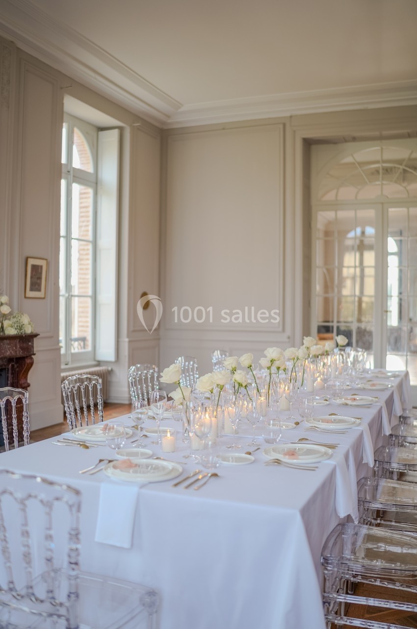 Table élégamment dressée avec nappes blanches, chaises transparentes, bougies et roses blanches dans une salle lumineuse.