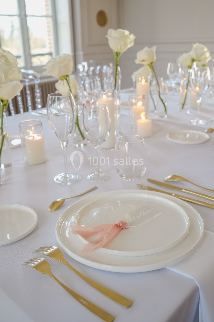 Table élégamment dressée avec vaisselle blanche, couverts dorés, bougies allumées et roses blanches en décoration.
