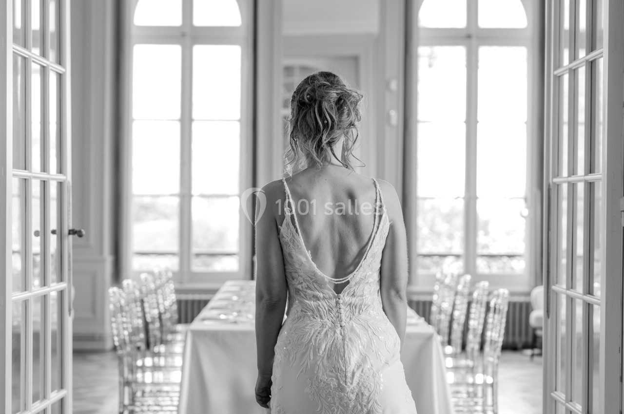 Une femme en robe de mariée vue de dos, debout dans une salle lumineuse avec de grandes fenêtres et une table dressée.