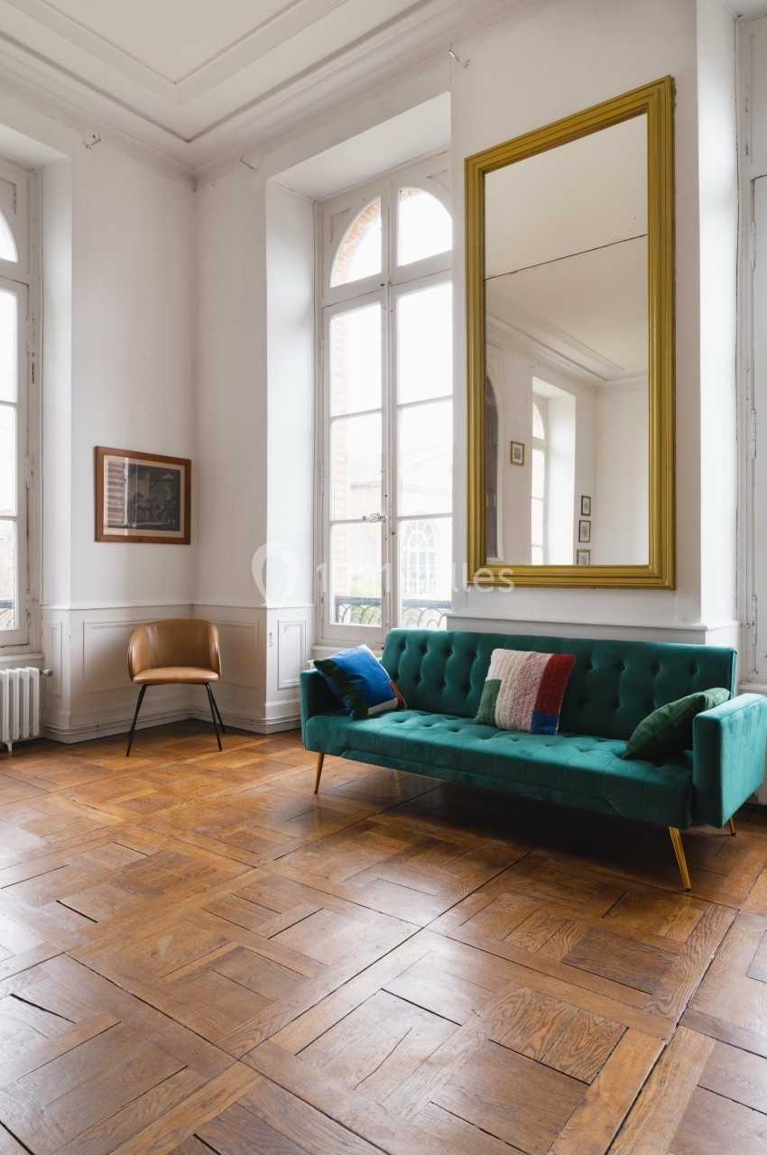 Salon lumineux avec parquet en bois, canapé vert, fauteuil beige, miroir doré et grandes fenêtres.