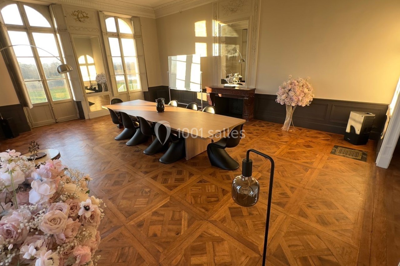 Salle lumineuse avec parquet en bois, grande table entourée de chaises noires, fleurs décoratives et fenêtres donnant sur l…