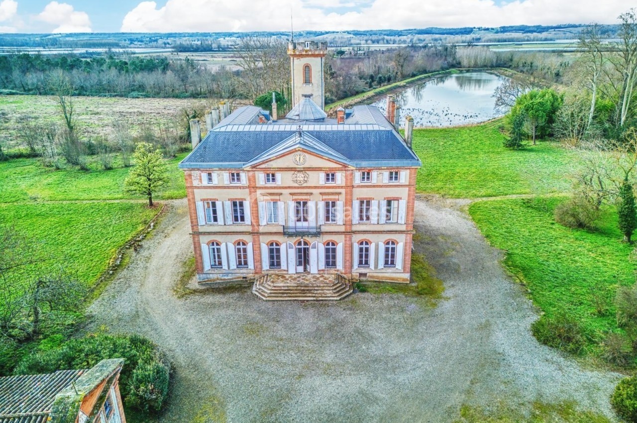 Vue aérienne d'un château en briques entouré de verdure, avec un étang en arrière-plan et un ciel partiellement nuageux.
