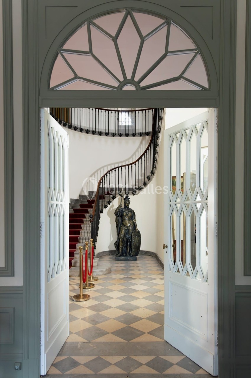Vue d'une entrée avec portes ouvertes sur un escalier en colimaçon, une statue et un sol en damier.