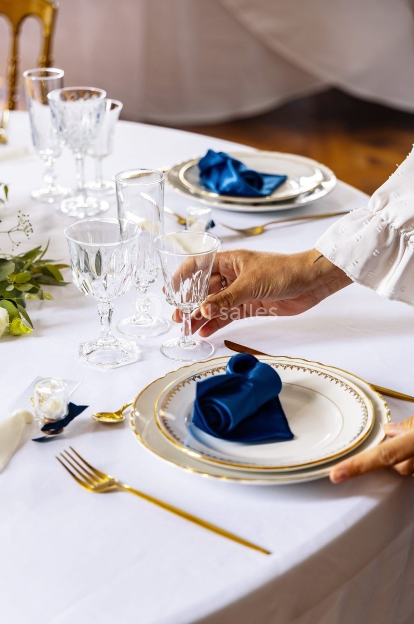 Une main dispose un verre en cristal sur une table élégamment dressée avec assiettes, couverts dorés et serviettes bleues.