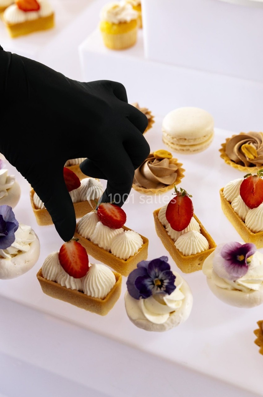 Main gantée disposant des pâtisseries décorées de fraises et fleurs comestibles sur une table blanche.