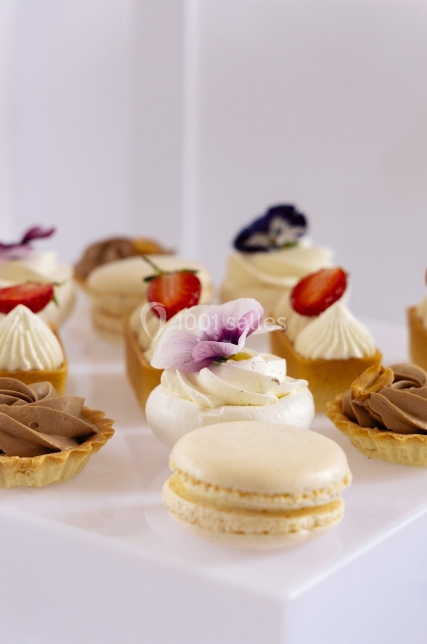 Assortiment de pâtisseries décorées de fleurs comestibles, fraises et crèmes, disposées sur une surface blanche.