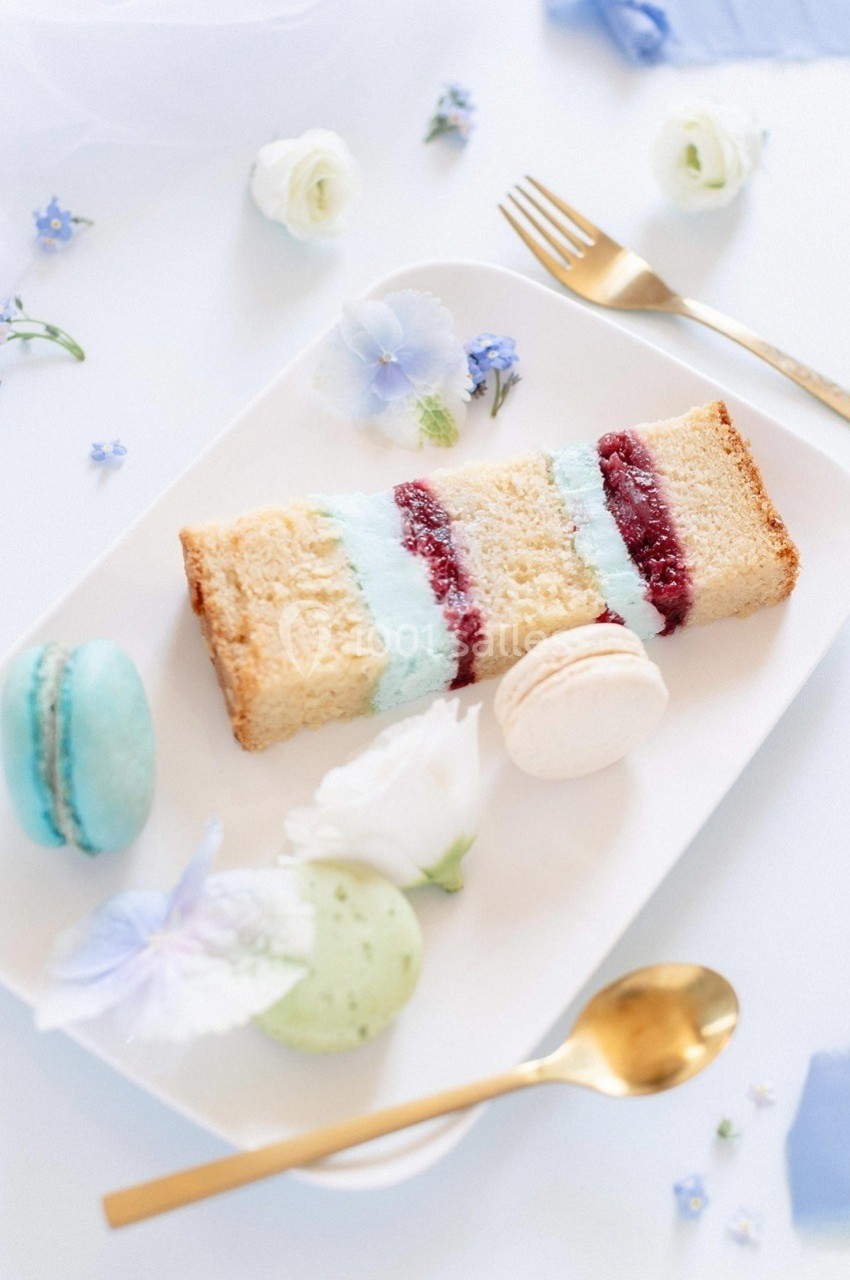 Assiette avec une part de gâteau à la crème colorée, macarons et fleurs décoratives sur une table blanche.