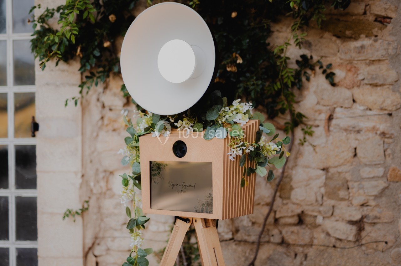 Appareil photo rétro en bois sur trépied, décoré de feuillage et fleurs, devant un mur en pierre.