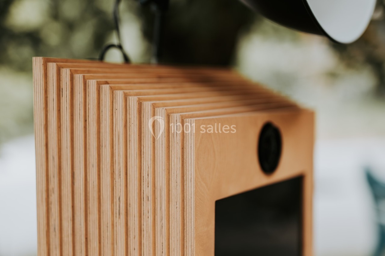 Boîtier en bois avec des rainures, équipé d'un écran et d'une caméra, placé en extérieur avec un fond flou.