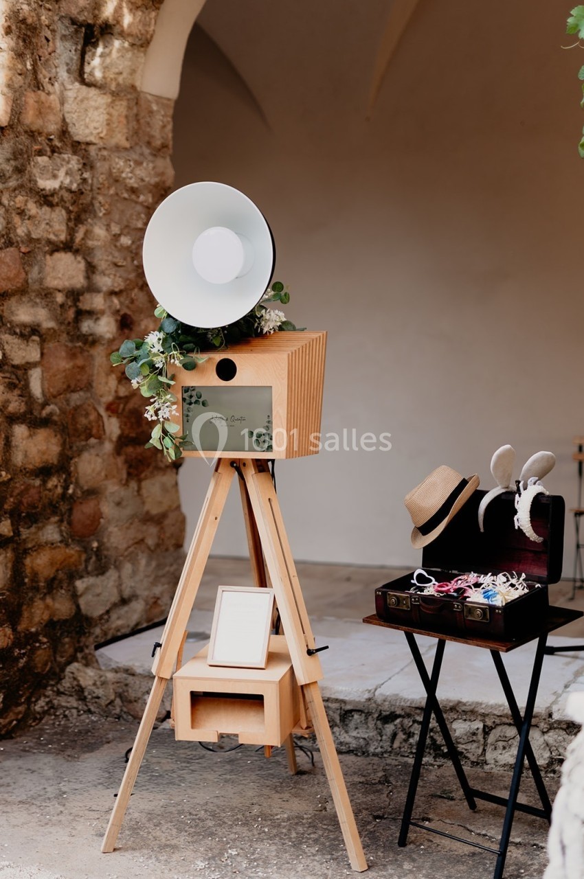 Photobooth en bois sur trépied avec accessoires dans une valise, installé devant un mur en pierre.