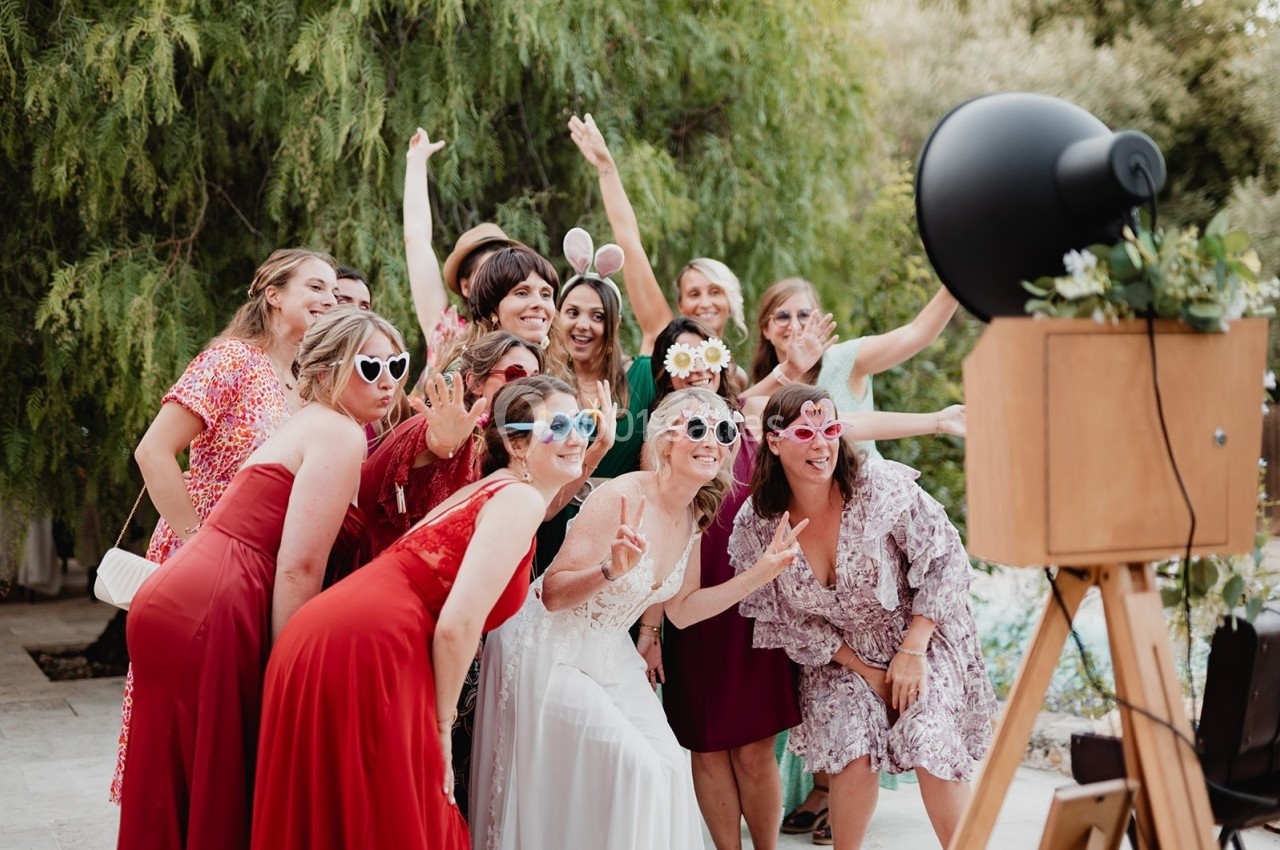 Un groupe de femmes en tenue festive pose avec des accessoires ludiques devant un photobooth en extérieur.