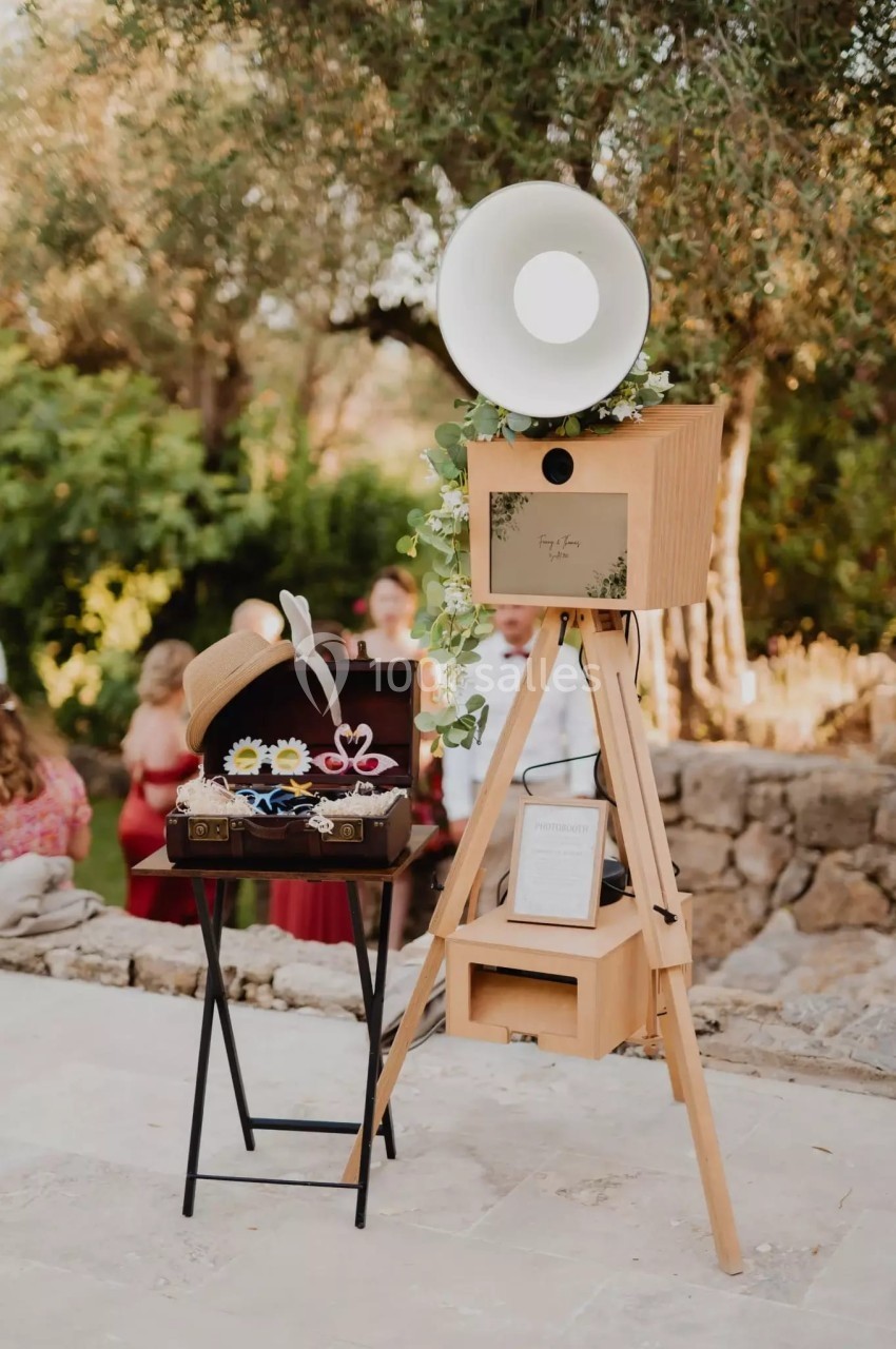 Photobooth en bois sur trépied avec accessoires festifs et décor champêtre en arrière-plan.