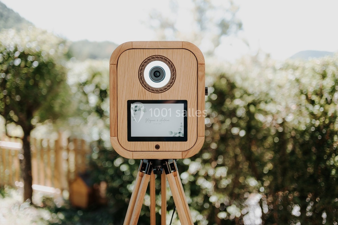 Appareil photo en bois sur trépied, placé dans un jardin avec des arbres et une clôture en arrière-plan.