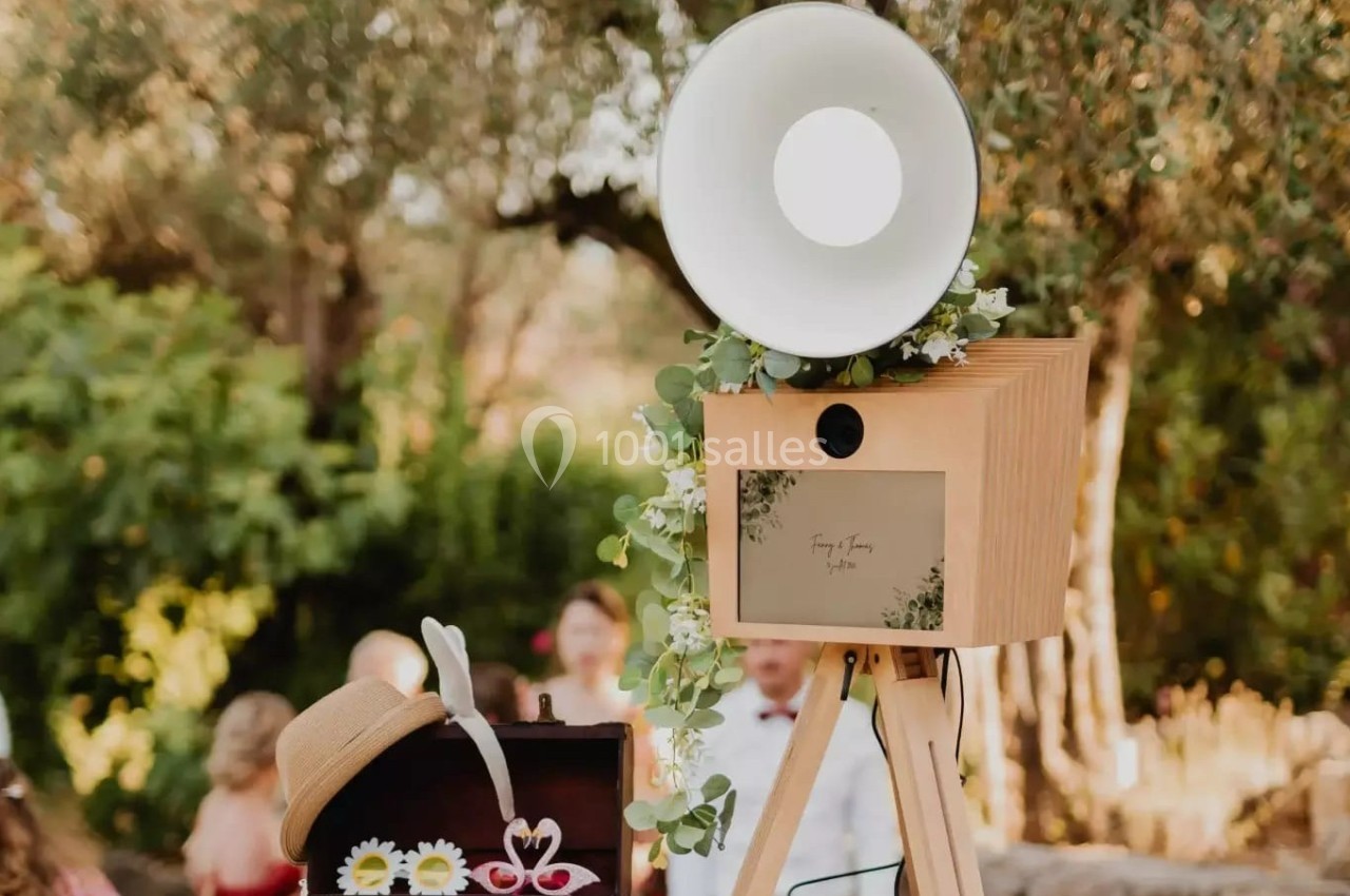 Photobooth en bois décoré de feuillage, avec une lampe circulaire, installé en extérieur lors d'un événement.