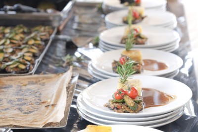 Un cuisinier en tenue noire prépare des légumes dans une poêle en extérieur, devant un mur en pierre.