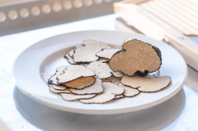 Un cuisinier en tenue noire prépare des légumes dans une poêle en extérieur, devant un mur en pierre.