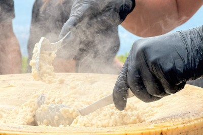 Un cuisinier en tenue noire prépare des légumes dans une poêle en extérieur, devant un mur en pierre.