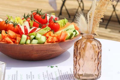 Un cuisinier en tenue noire prépare des légumes dans une poêle en extérieur, devant un mur en pierre.