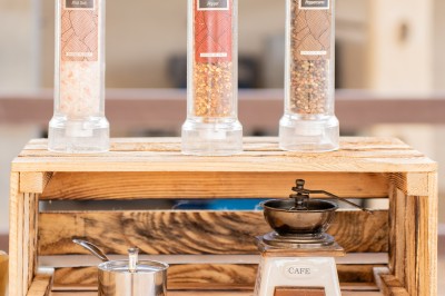 Un cuisinier en tenue noire prépare des légumes dans une poêle en extérieur, devant un mur en pierre.