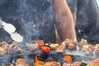 Un cuisinier en tenue noire prépare des légumes dans une poêle en extérieur, devant un mur en pierre.