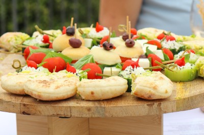 Un cuisinier en tenue noire prépare des légumes dans une poêle en extérieur, devant un mur en pierre.