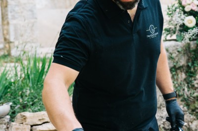 Un cuisinier en tenue noire prépare des légumes dans une poêle en extérieur, devant un mur en pierre.