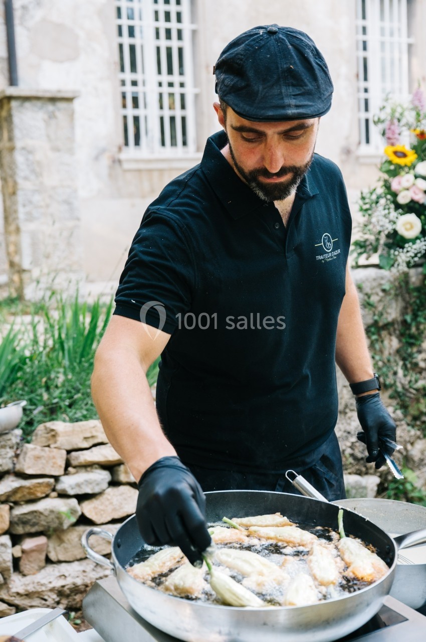 Un cuisinier en tenue noire prépare des légumes dans une poêle en extérieur, devant un mur en pierre.