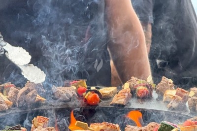 Viande et légumes grillés sur une plancha avec des flammes, cuisinés par une personne en polo noir.