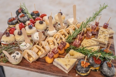 Plateau en bois garni de bouchées variées, incluant mini-sandwichs, verrines, fromages et décorations de romarin.