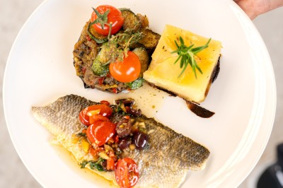 Un cuisinier en tenue noire prépare des légumes dans une grande poêle en extérieur, devant un mur en pierre.