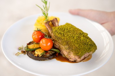 Un cuisinier en tenue noire prépare des légumes dans une grande poêle en extérieur, devant un mur en pierre.