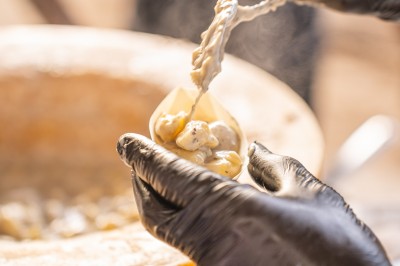 Un cuisinier en tenue noire prépare des légumes dans une grande poêle en extérieur, devant un mur en pierre.