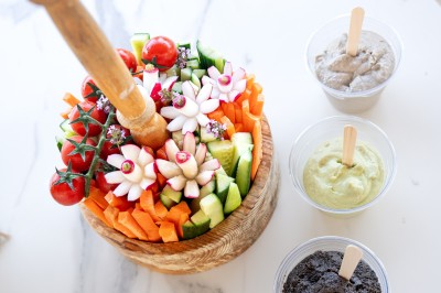 Un cuisinier en tenue noire prépare des légumes dans une grande poêle en extérieur, devant un mur en pierre.