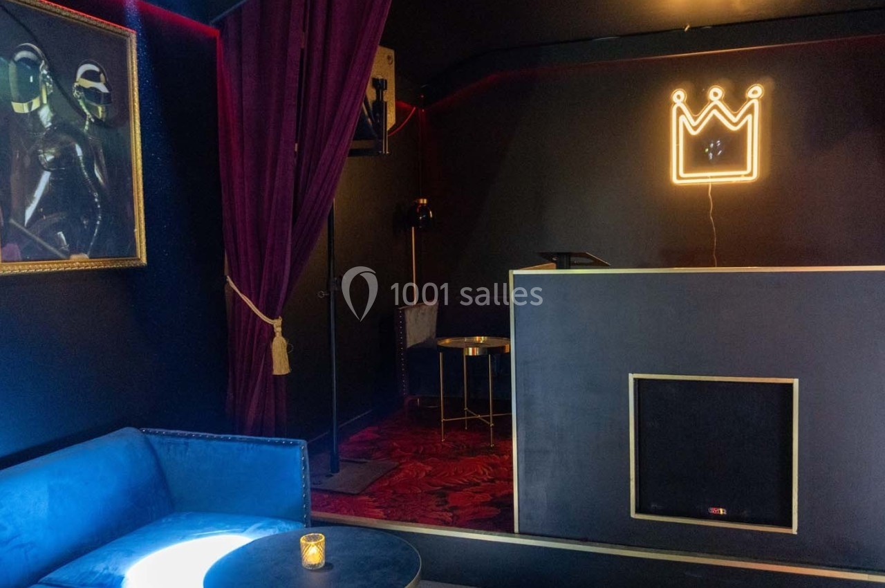 Salon sombre avec canapé bleu, table basse, rideaux pourpres, tapis rouge et enseigne lumineuse en forme de couronne.