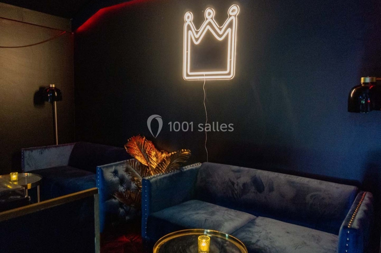 Salon sombre avec canapés bleus, table basse, lampe et décoration murale en néon représentant une couronne.