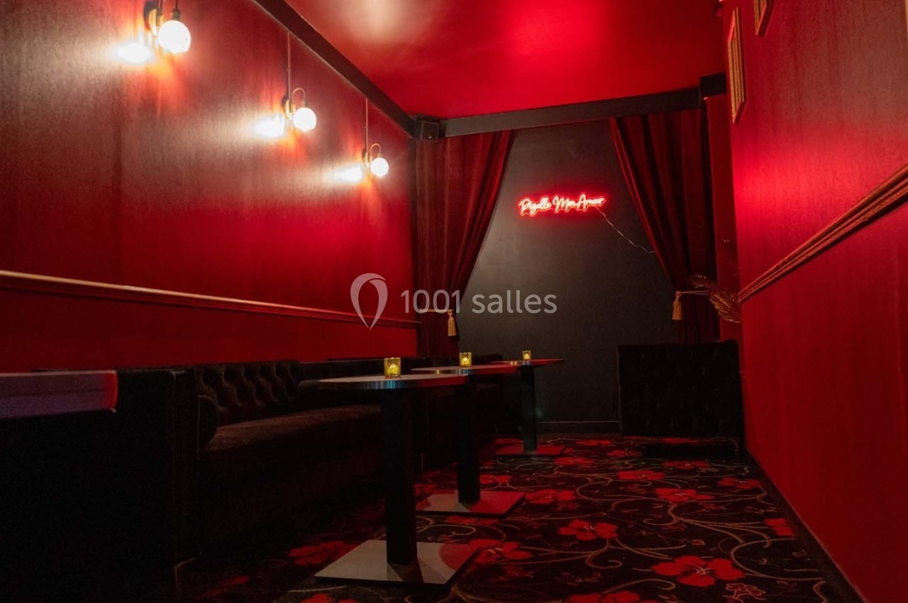 Salle rouge avec banquettes noires, tables sombres, tapis à motifs et enseigne lumineuse au mur.