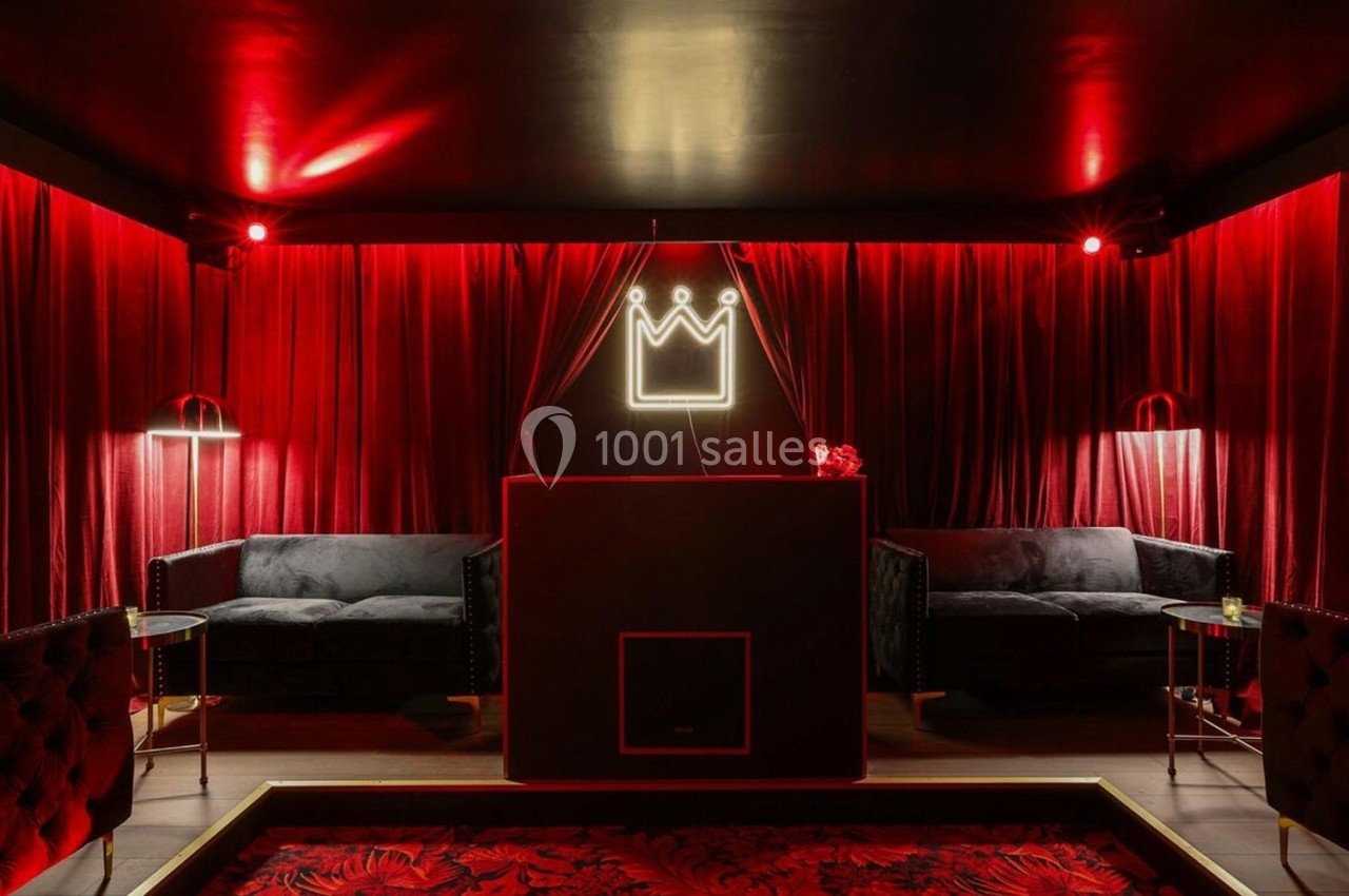 Salle sombre avec rideaux rouges, canapé noir, éclairage tamisé et enseigne lumineuse en forme de couronne.