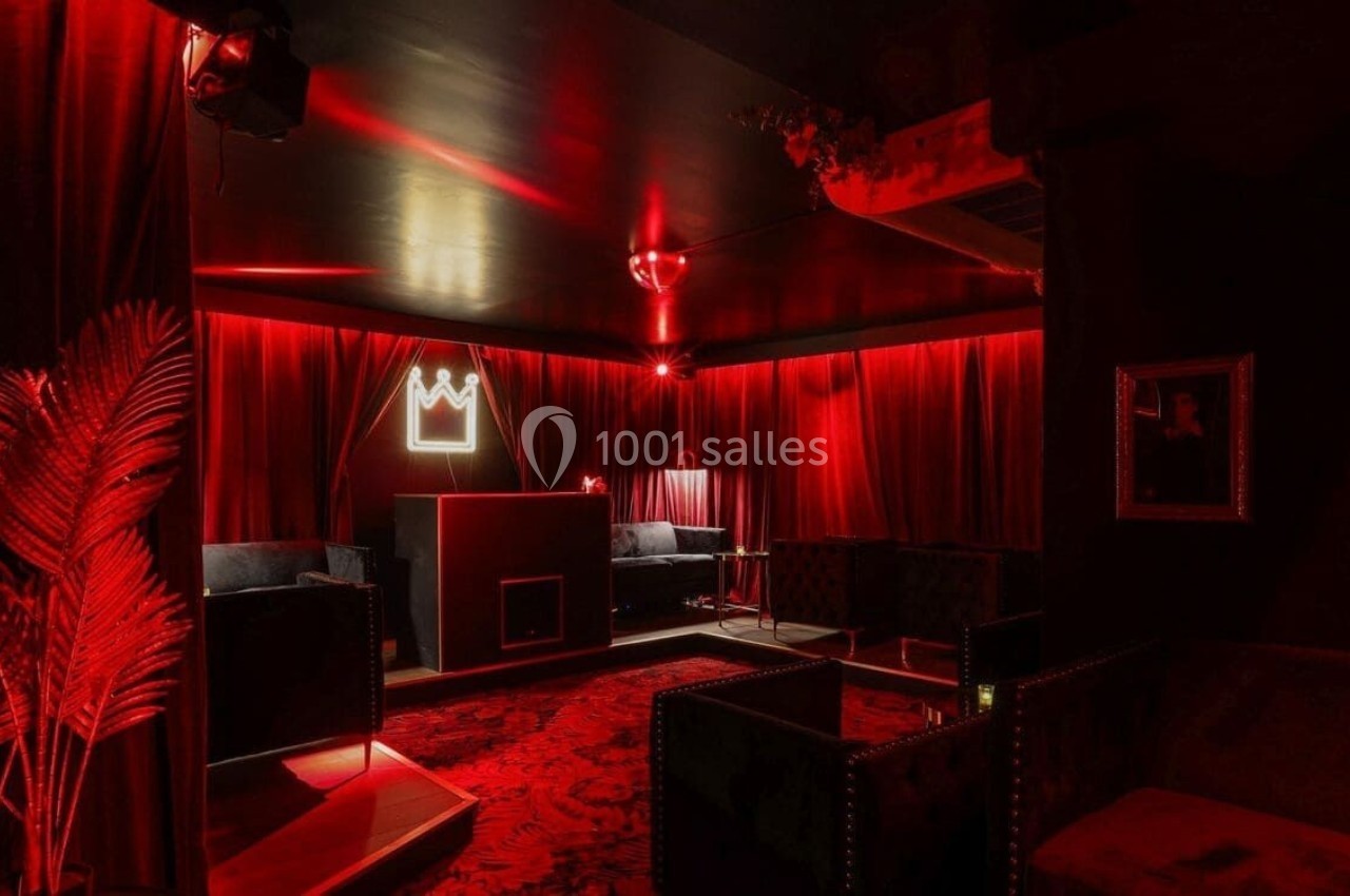 Salle sombre avec éclairage rouge, rideaux en velours, canapés noirs et un néon en forme de couronne sur le mur.