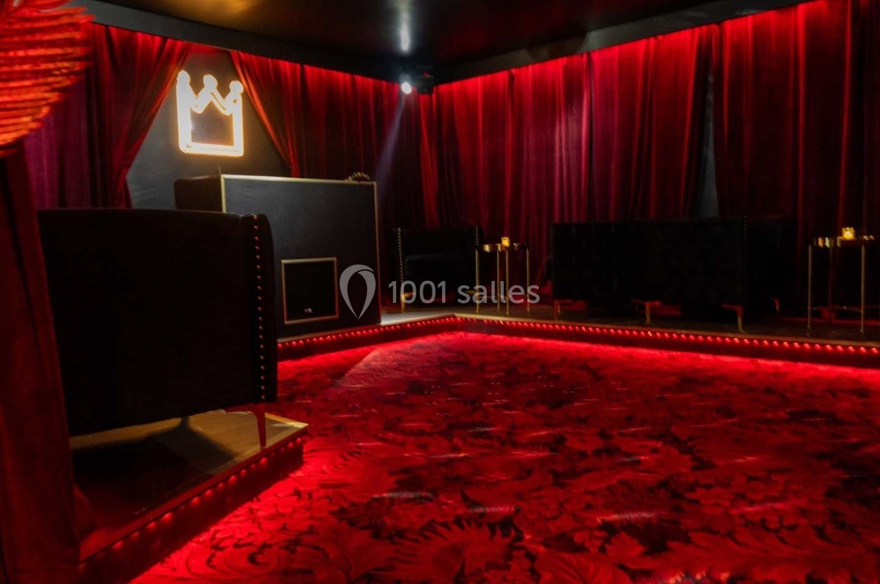 Salle sombre avec rideaux rouges, éclairage tamisé, tapis rouge orné et mobilier noir, ambiance intime et élégante.