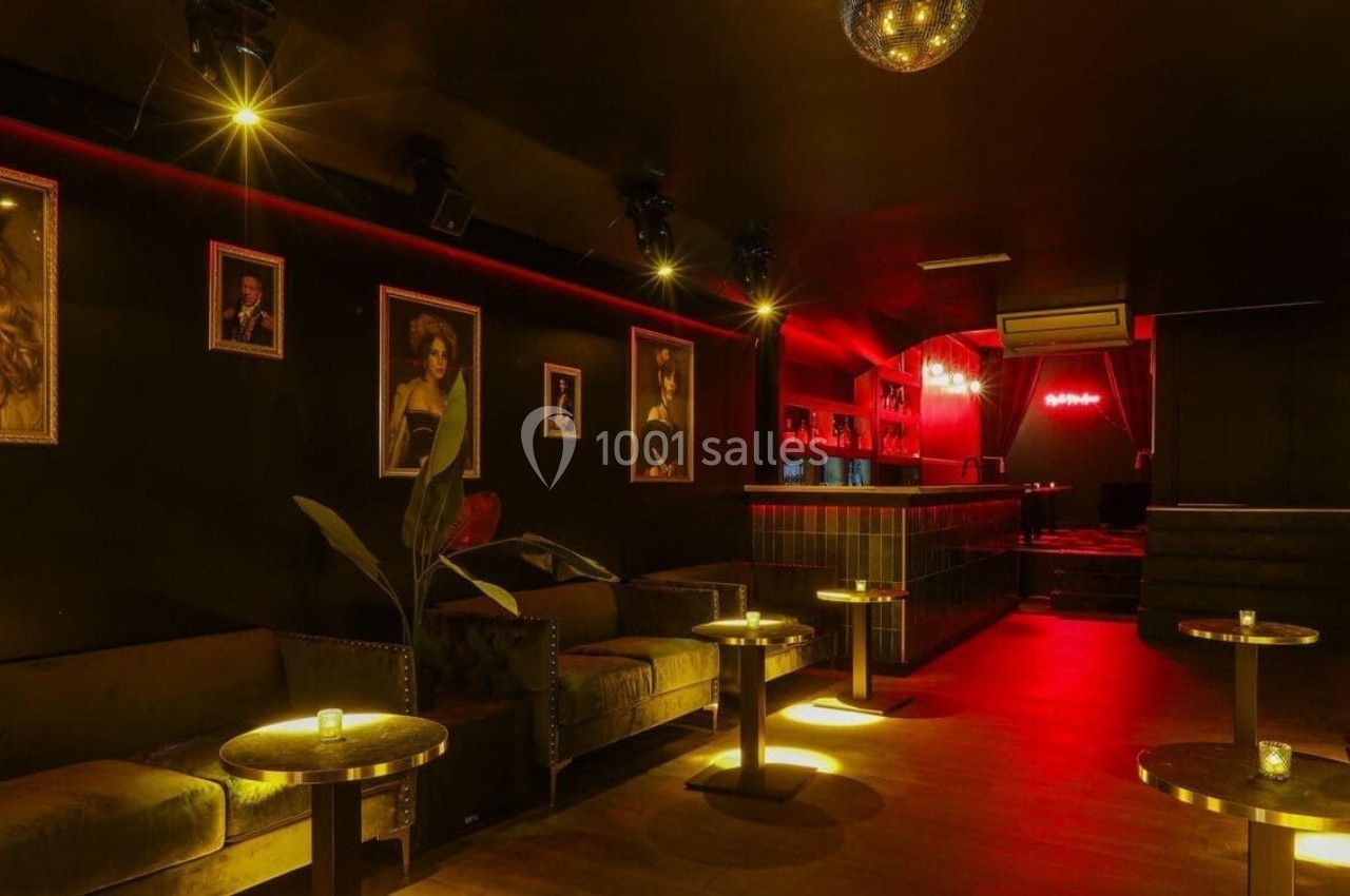 Salle de bar tamisée avec canapés, tables dorées, portraits encadrés, éclairage rouge et boule disco au plafond.